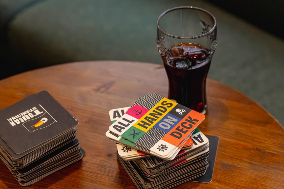 A wooden pub table with two stacks of branded beer mats and a glass of Coke.