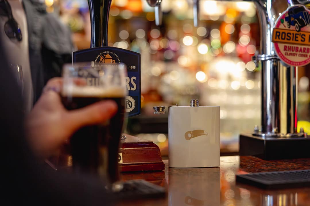 A traditional pub bar with beer pumps and a Toucan branded white hip flask with a Toucan's head engraved into it.