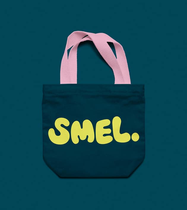 Dark blue SMEL tote bag with soft pink handles, placed on a deep teal background. The bag is printed with the bold yellow SMEL logo in playful, rounded lettering across the front.