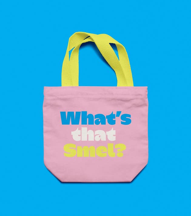 Light pink SMEL tote bag with neon yellow handles, shown against a bright blue background. The front of the bag features the phrase "What's that Smel?" in large, colourful block lettering in blue, white, and yellow.