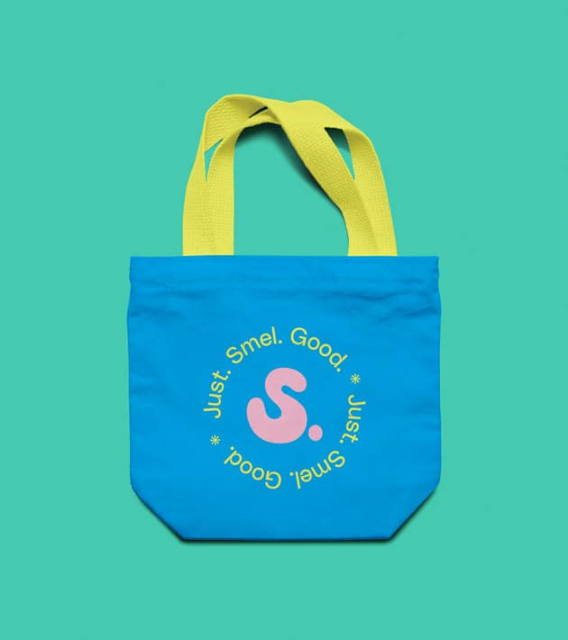 Bright blue SMEL tote bag with neon yellow handles, displayed against a turquoise background. The design features a large pink "S." surrounded by the phrase "Just. Smel. Good." repeated in a circle in yellow text.