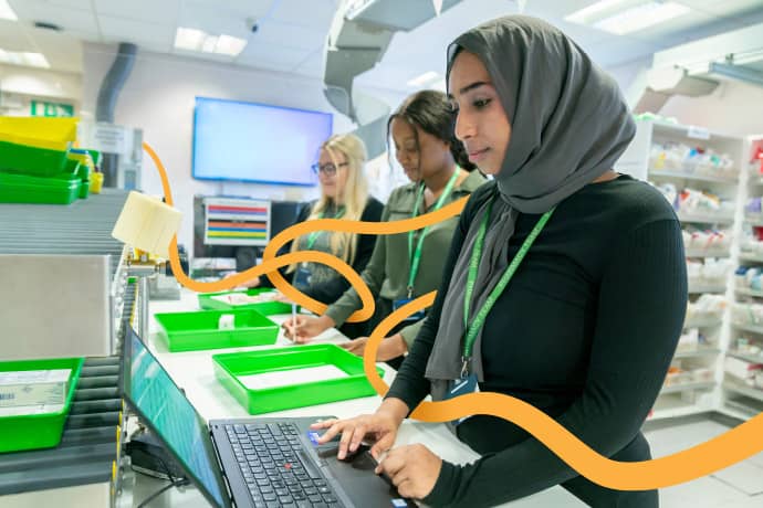 3 young female pharmacists at a counter. One is on a laptop, the other 2 are writing.