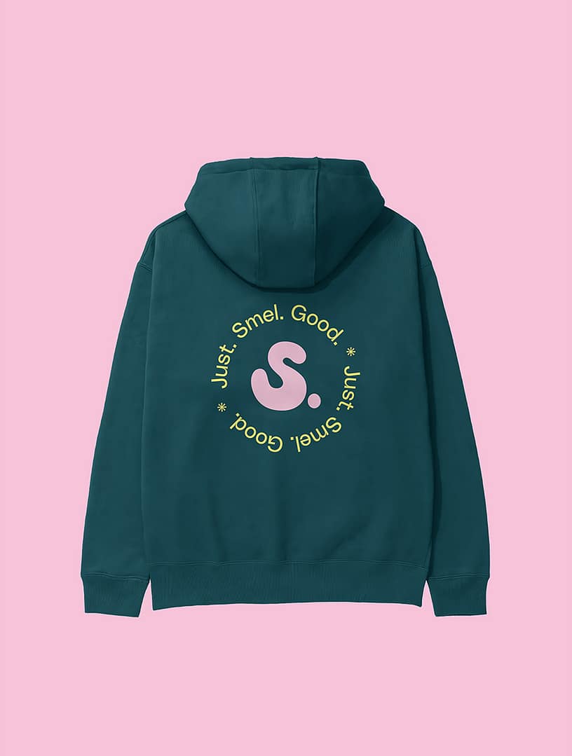 Back view of a dark teal SMEL hoodie against a soft pink background. The design features a large circular graphic with the phrase "Just. Smel. Good." repeated around a central pale pink "S." logo.