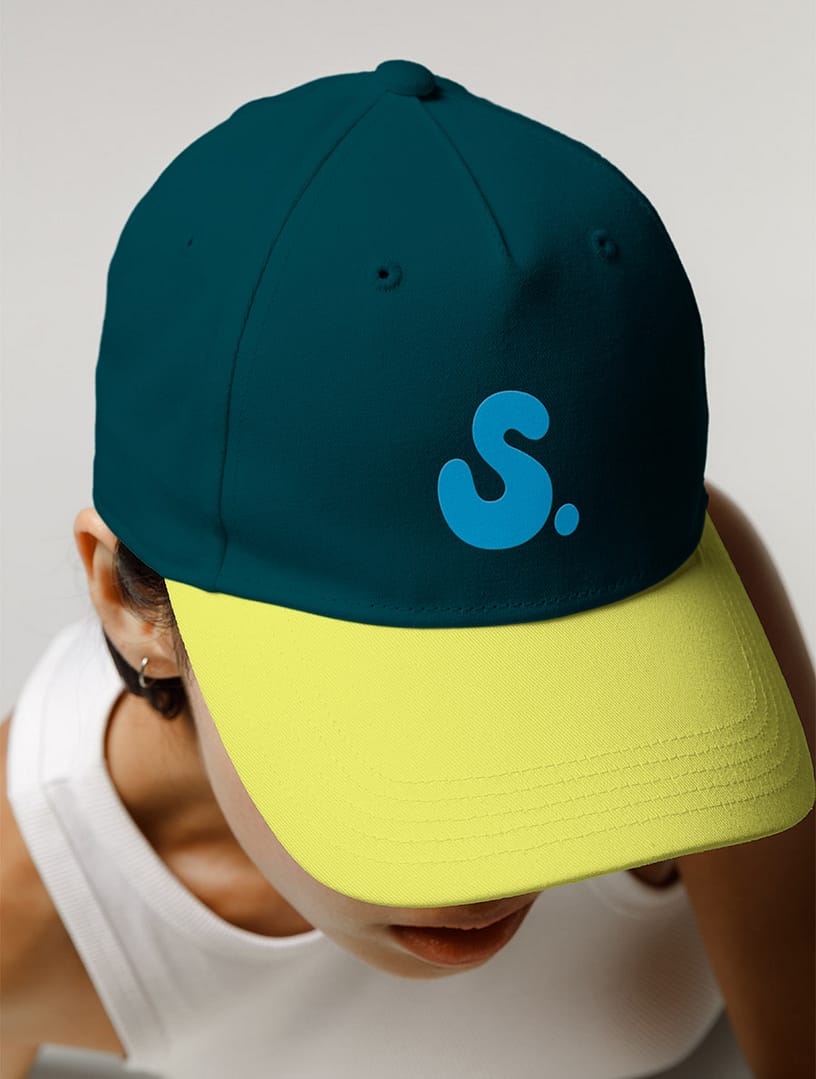 Close-up of a person wearing a two-tone SMEL baseball cap with a dark teal crown and bright yellow brim. A bold blue "S." logo is printed on the front. The wearer is dressed in a white top, with the background kept neutral.